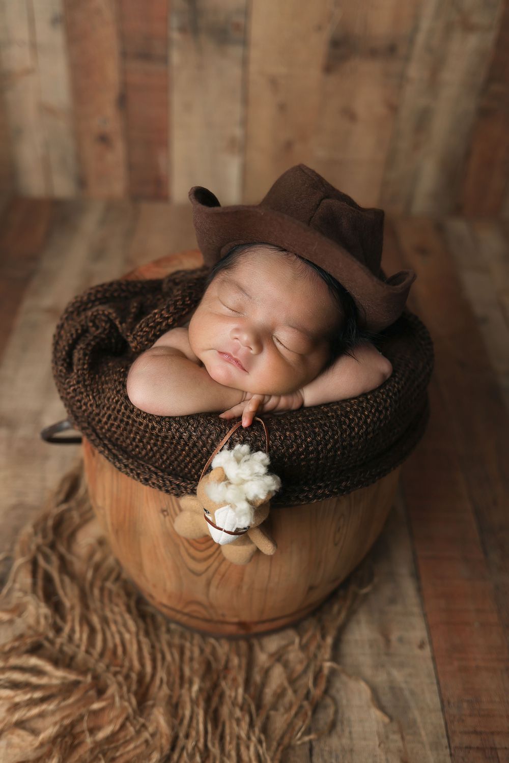 A sleeping newborn rests peacefully in a brown knit blanket inside a wooden bowl with cotton flowers.