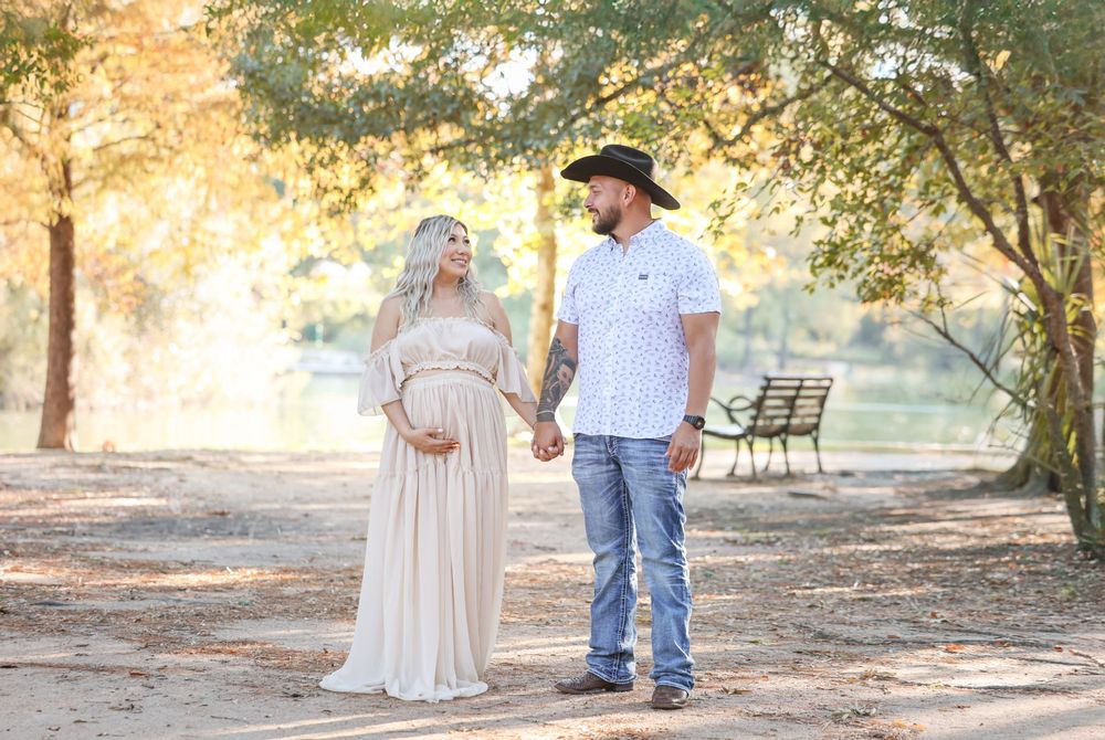A couple holds hands on a sunlit path in a park, with the person on the left wearing a flowing white maternity dress.