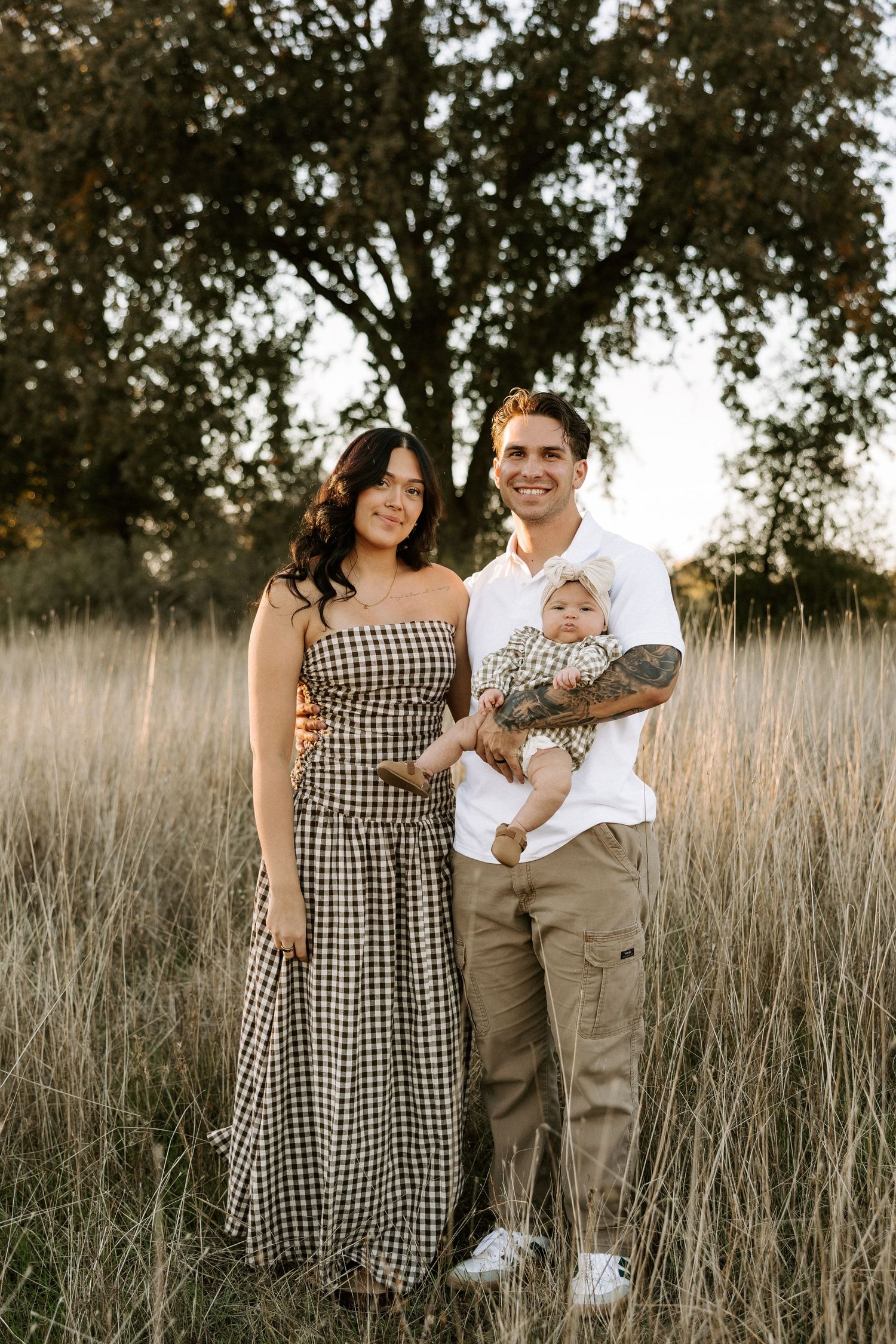 A couple and baby pose together in a field of tall grass at sunset with trees in the background.