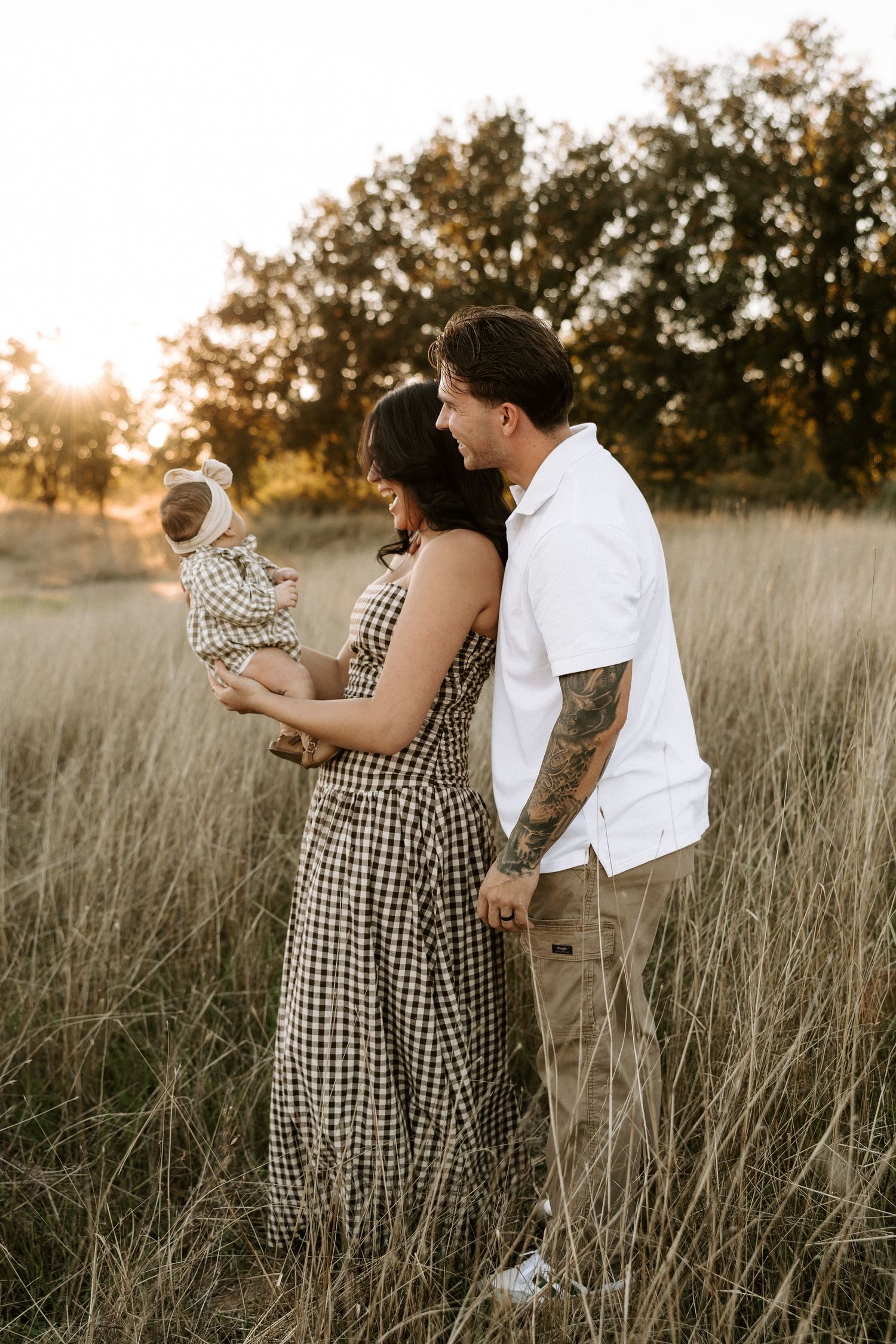 A couple and their baby stand together in a golden sunlit field of tall grass at sunset.