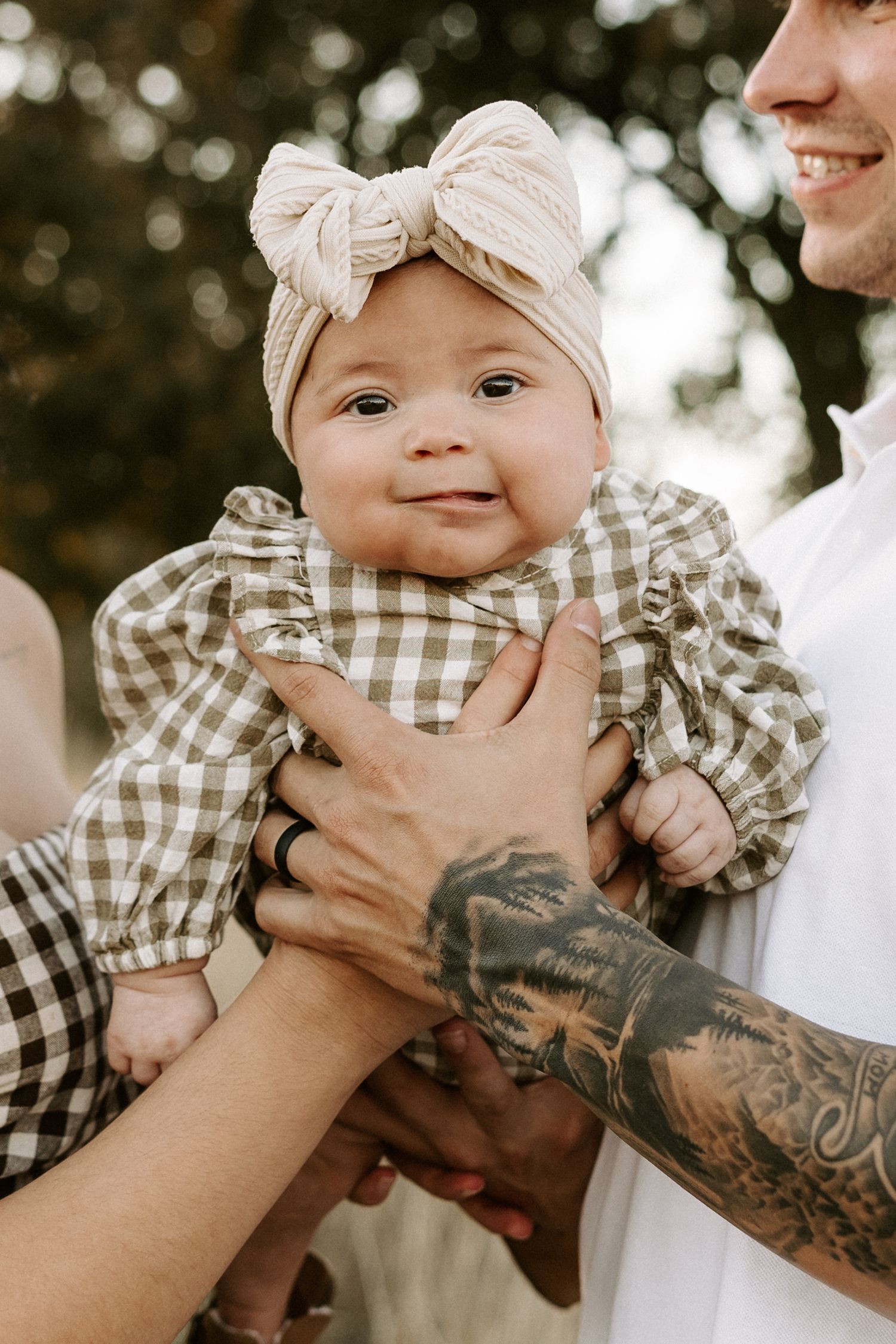 A series of four photos showing a baby in gingham clothing being held by tattooed arms.