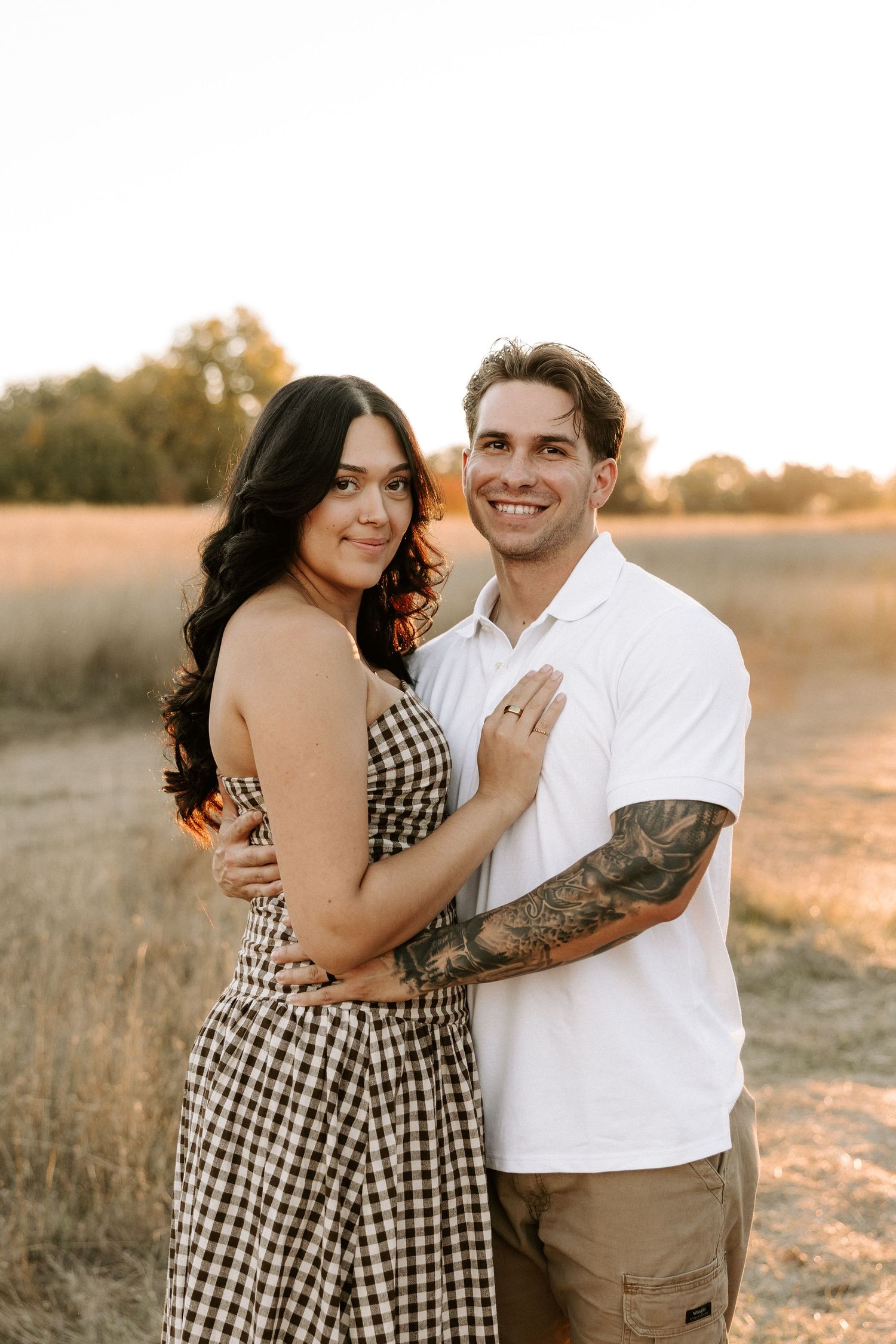 A couple embraces in a field at golden hour wearing a gingham dress and white shirt with khaki pants.