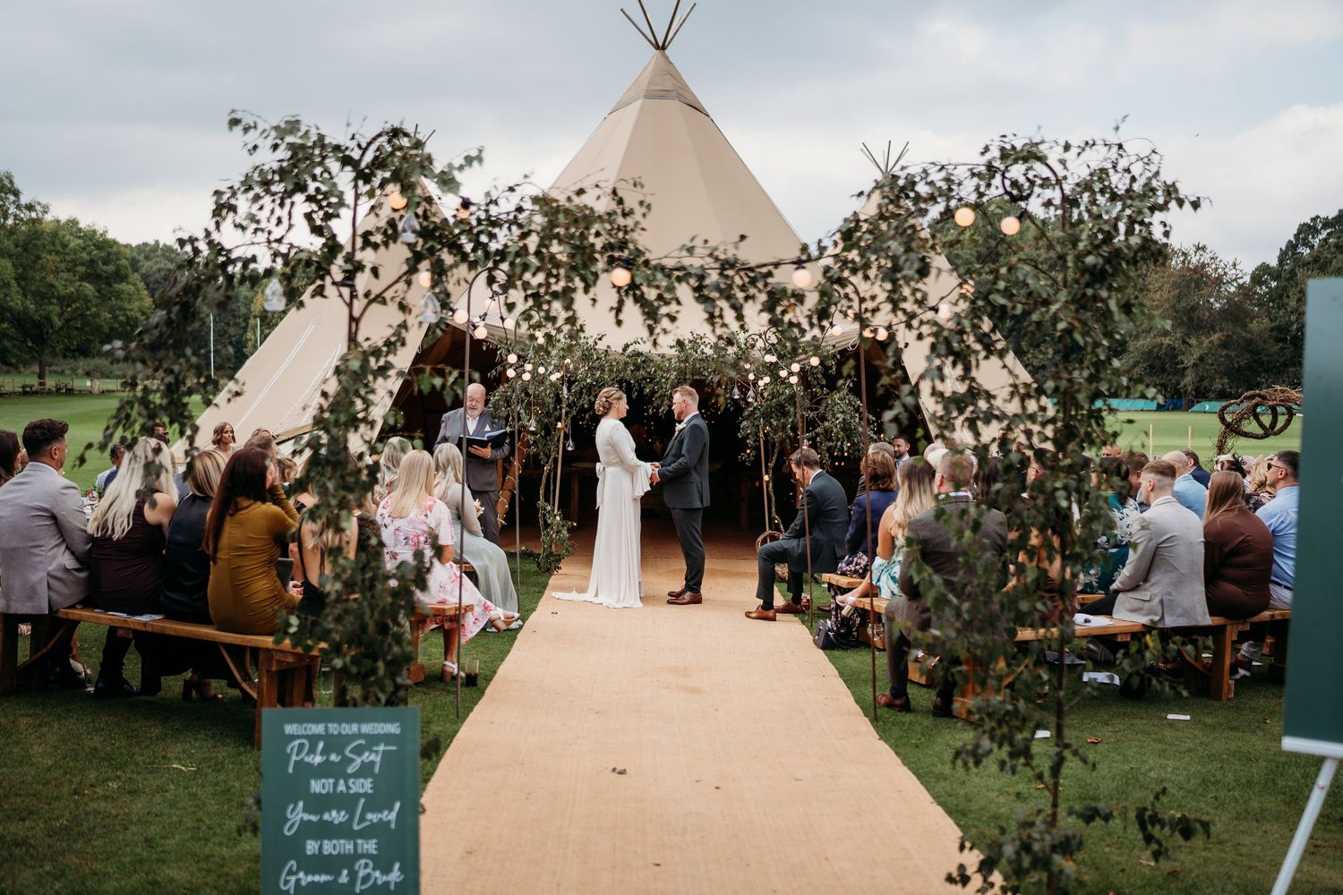 A wedding ceremony outside a tipi tent decorated with greenery and wooden chairs.