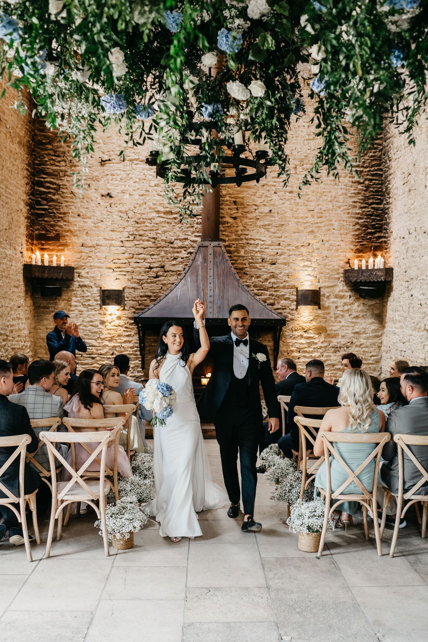 Elegant wedding ceremony in rustic stone venue with greenery-draped chandelier and cross-back chairs lining the aisle.