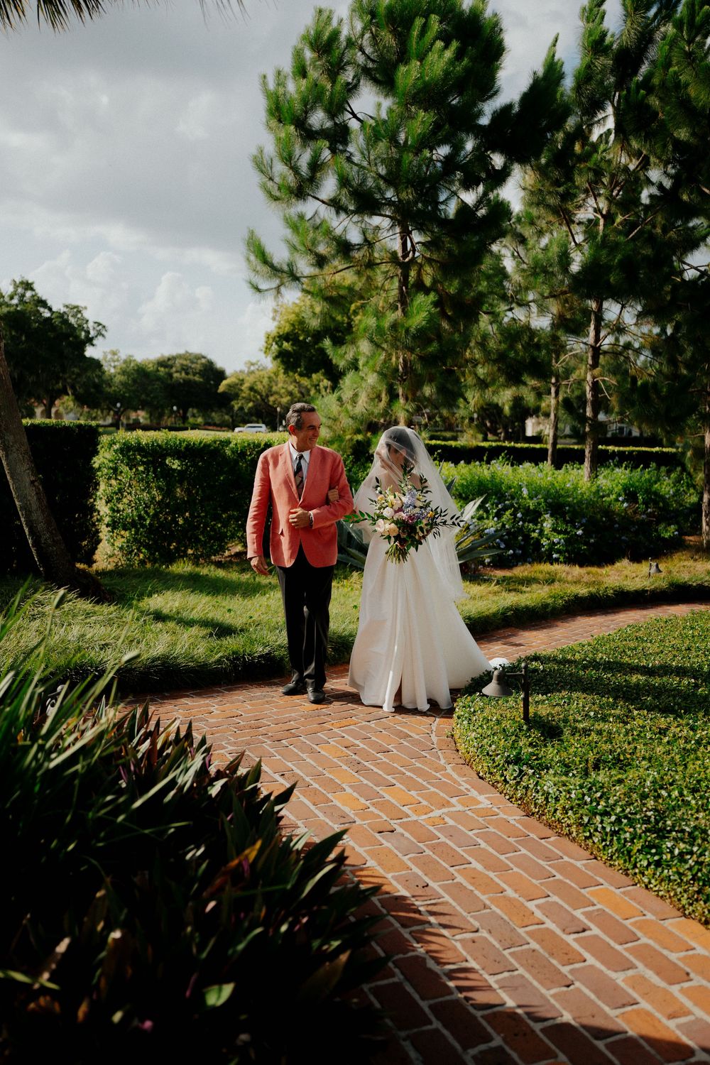 Wedding couple strolling down a winding brick pathway surrounded by lush garden landscaping.