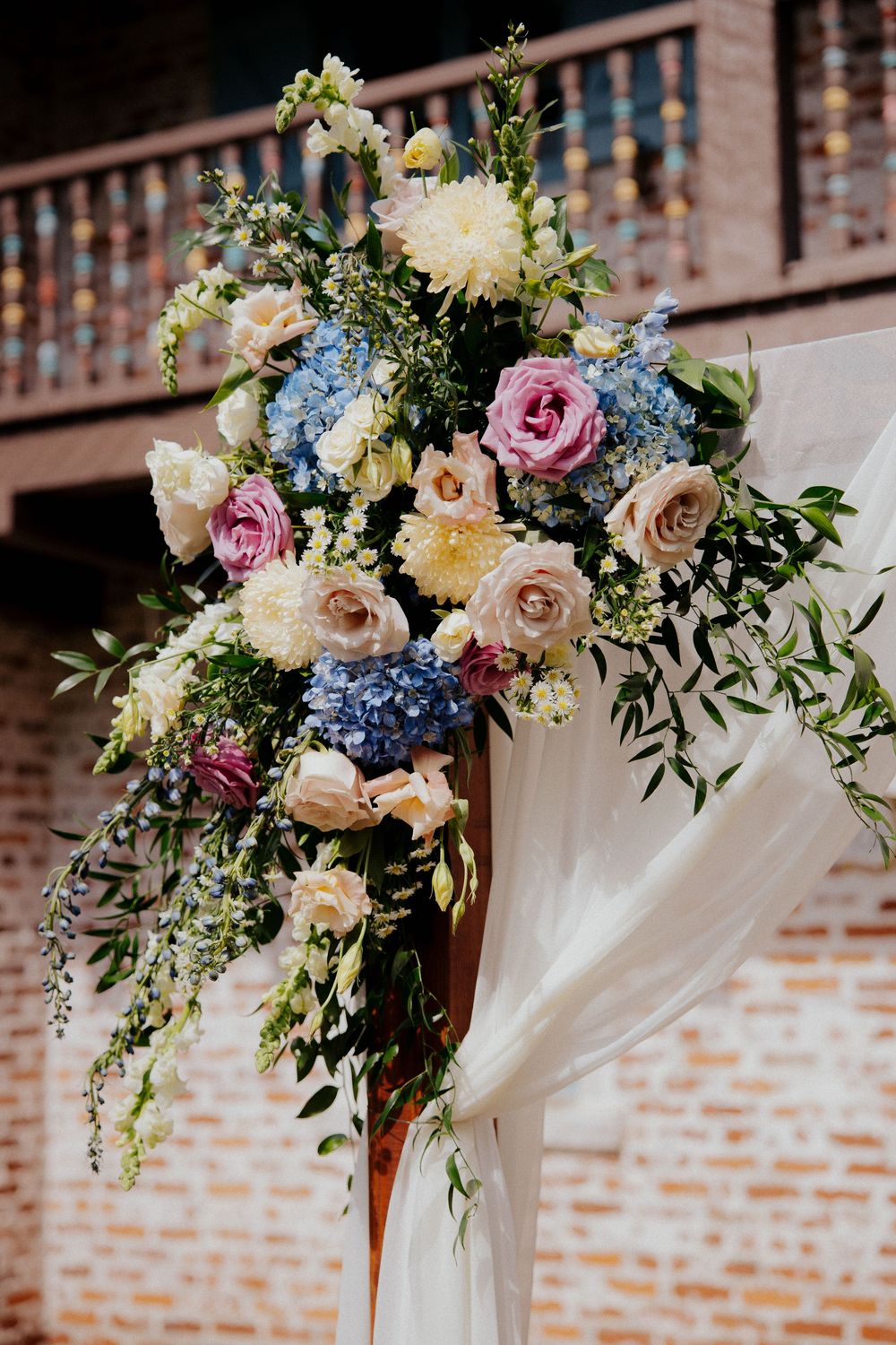 Floral wedding arch with pink roses, blue hydrangeas and white draping against a brick wall.