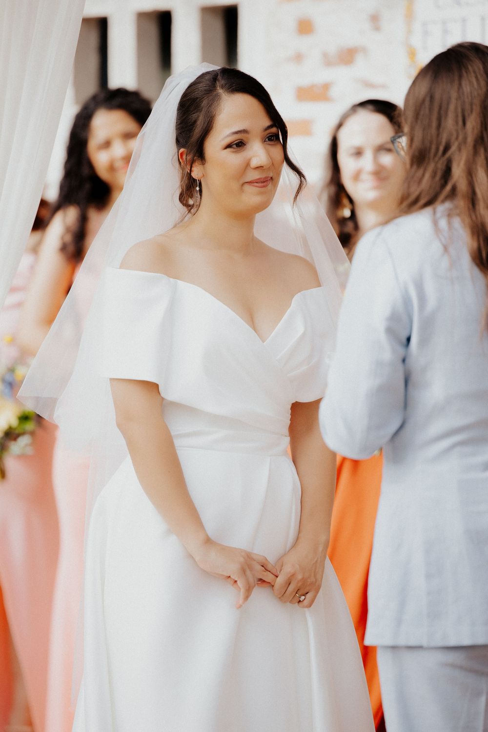 A bride in an elegant off-shoulder white wedding dress stands during a ceremony with bridesmaids in pastel dresses behind her.