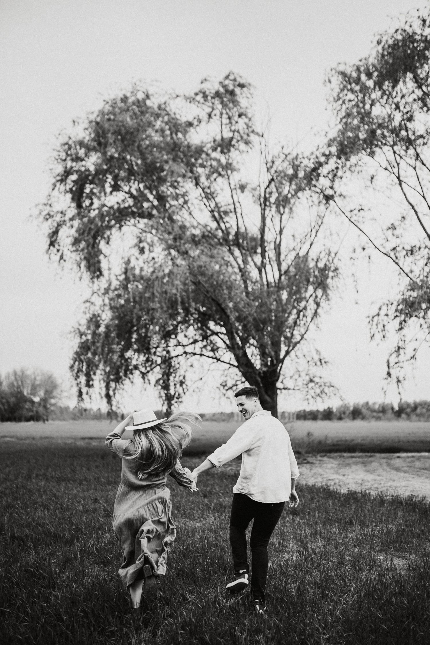 Playful couple running through field with willow tree in background.