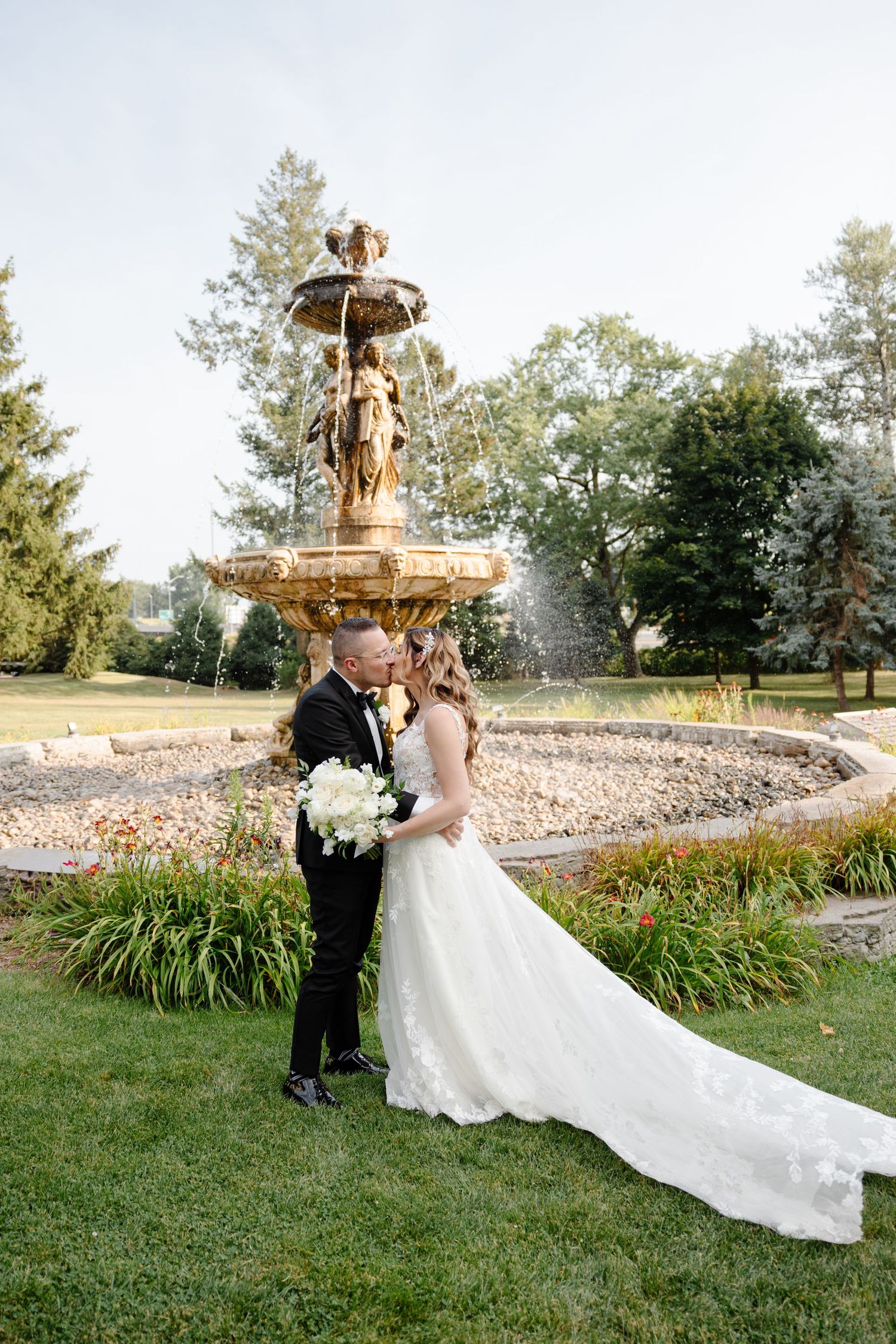A couple in formal attire poses near a decorative water fountain in a garden setting during golden hour.