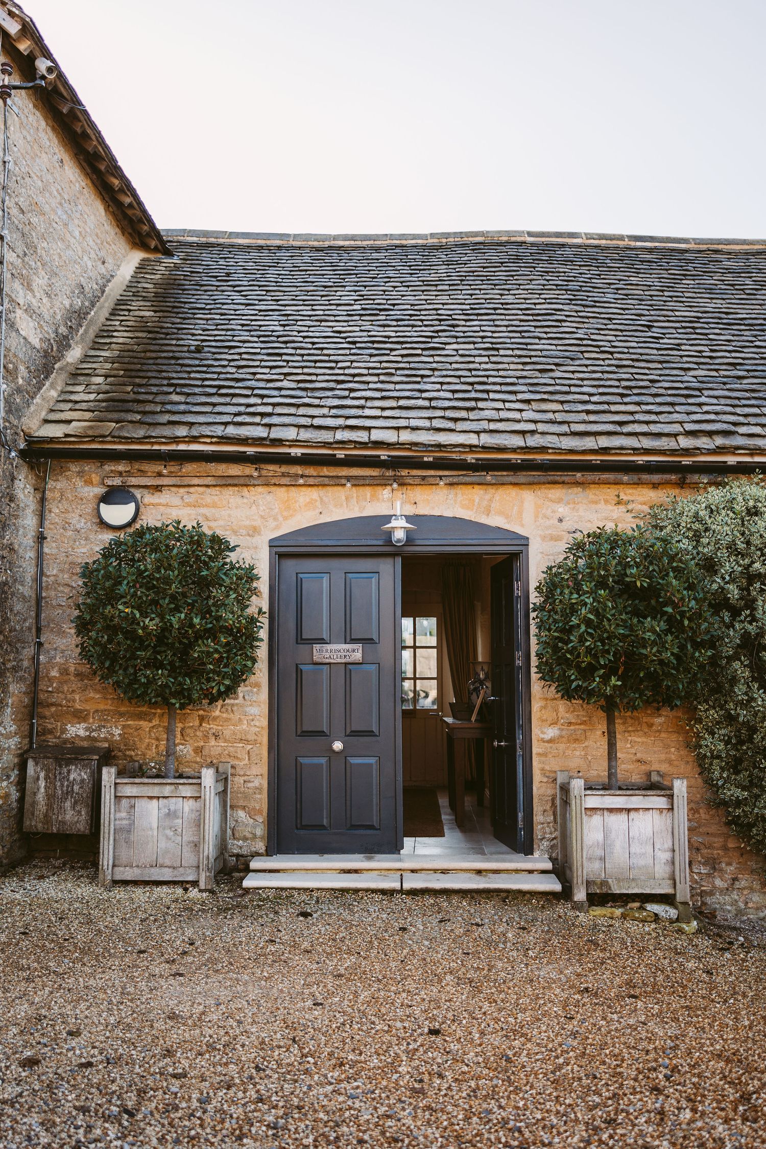 Stone cottage entrance with black doors, gravel courtyard, and potted topiary plants flanking the doorway.
