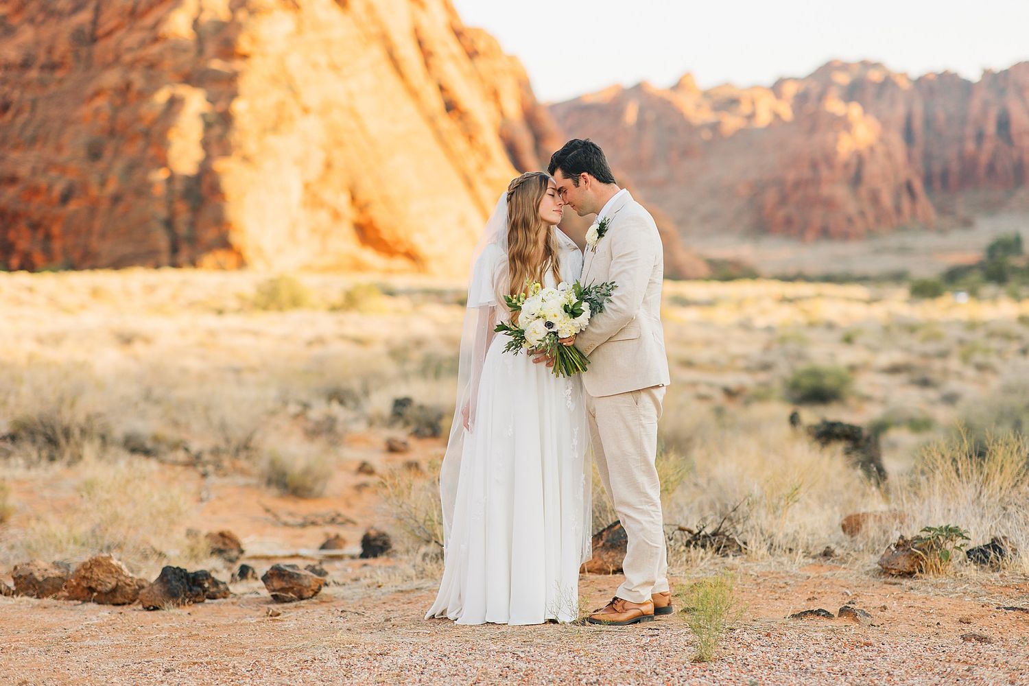 A romantic wedding couple embraces in a desert landscape with red rock formations at sunset.