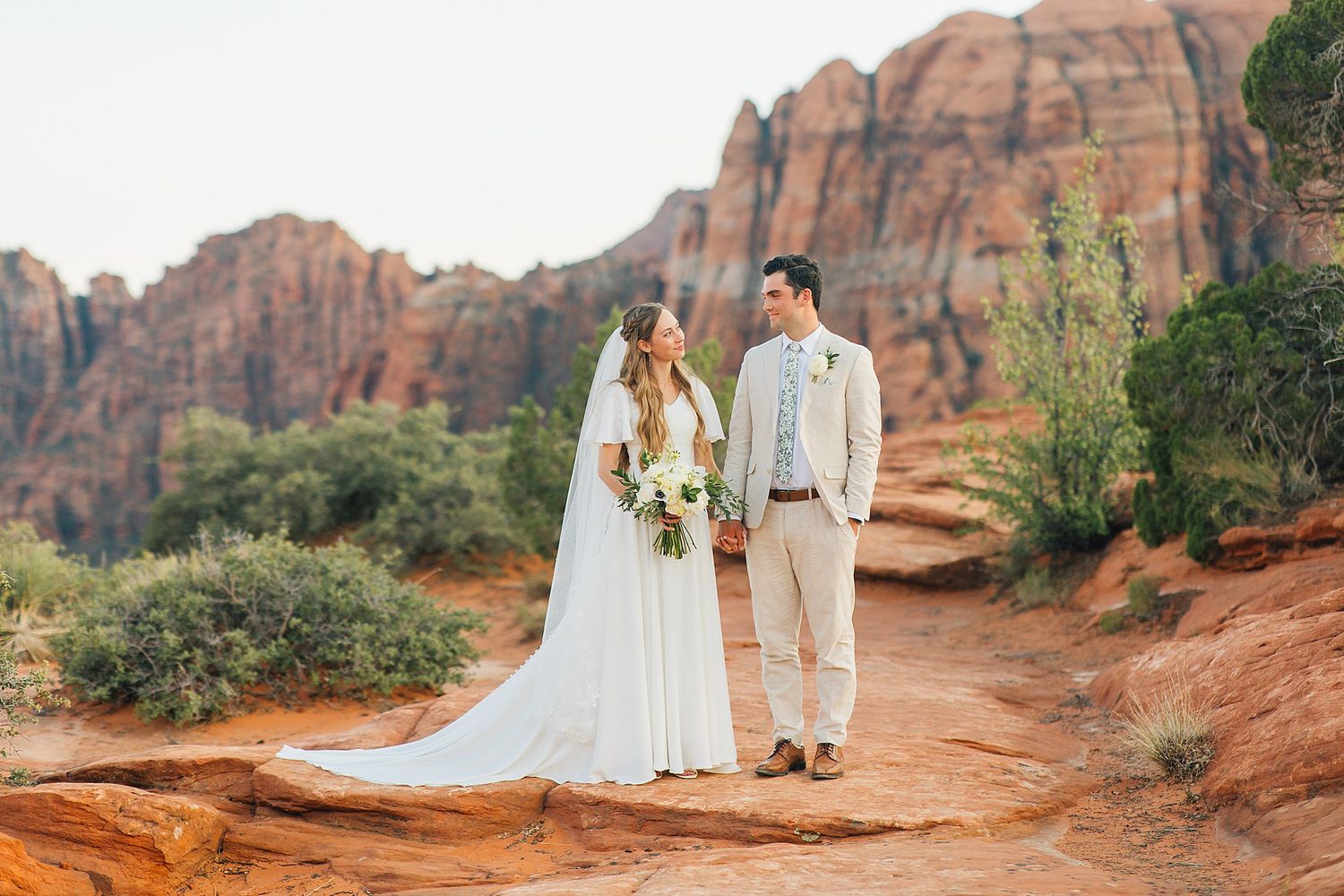 A couple in wedding attire poses together against stunning red rock formations in Sedona during sunset.