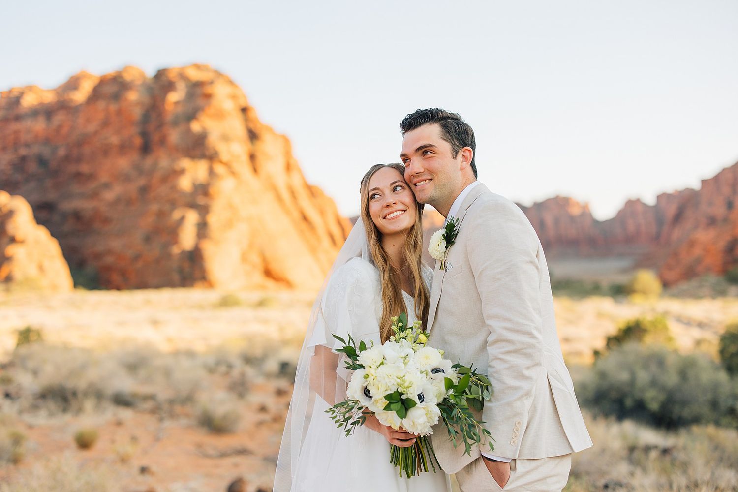 Newlyweds share intimate moments among desert landscape with dramatic sandstone cliffs in background.
