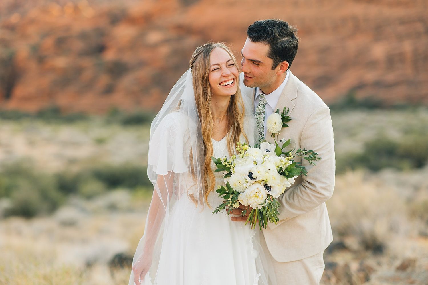 A couple shares a romantic moment in a desert landscape with red rock formations in the background.
