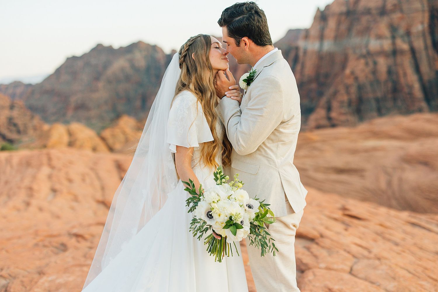 Newlywed couple shares intimate moment against stunning backdrop of desert cliffs and red rocks.