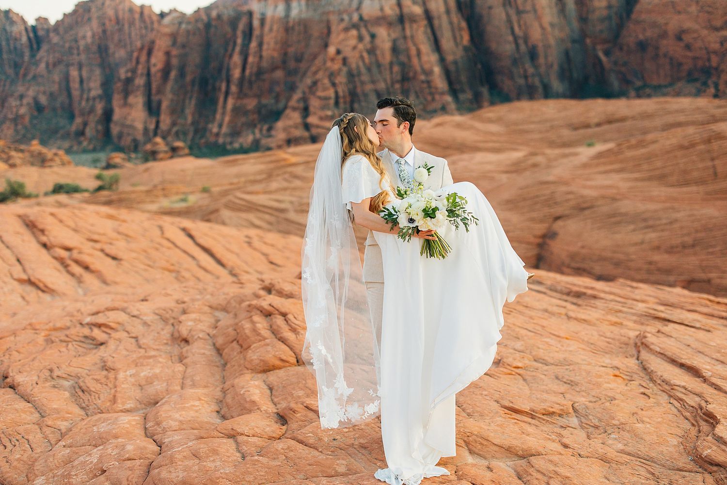 Couple in wedding attire stands on red rock formations at sunset in a desert landscape with dramatic cliffs.