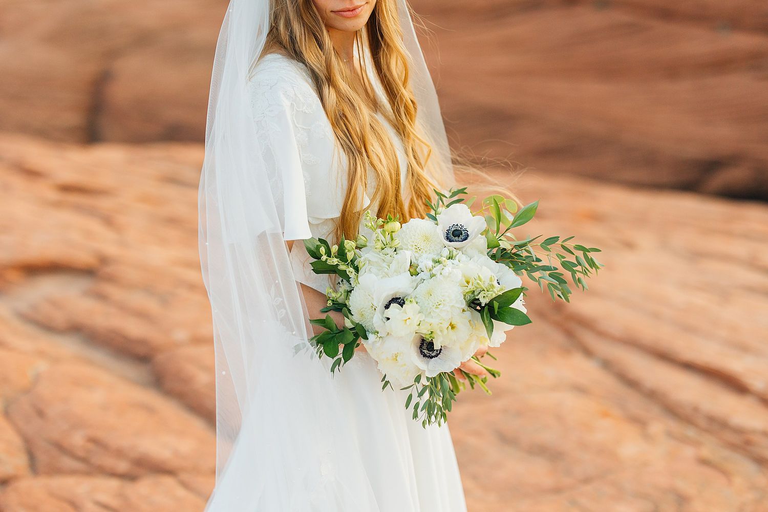 A bride in a white dress holds a delicate bouquet of white and green flowers against a desert sandstone backdrop.