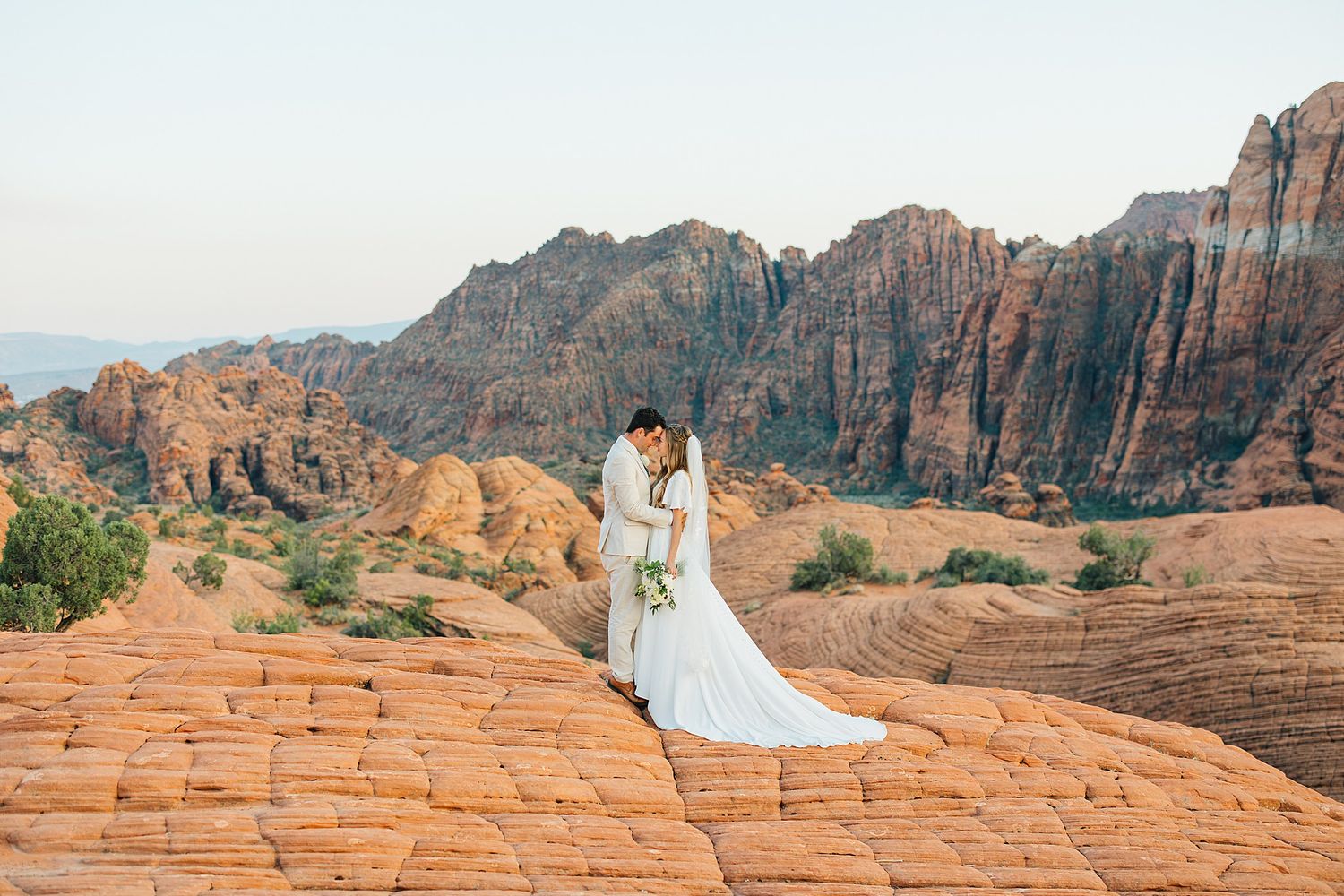 Newlyweds embrace on red sandstone cliffs with sweeping desert mountain vistas in the background.
