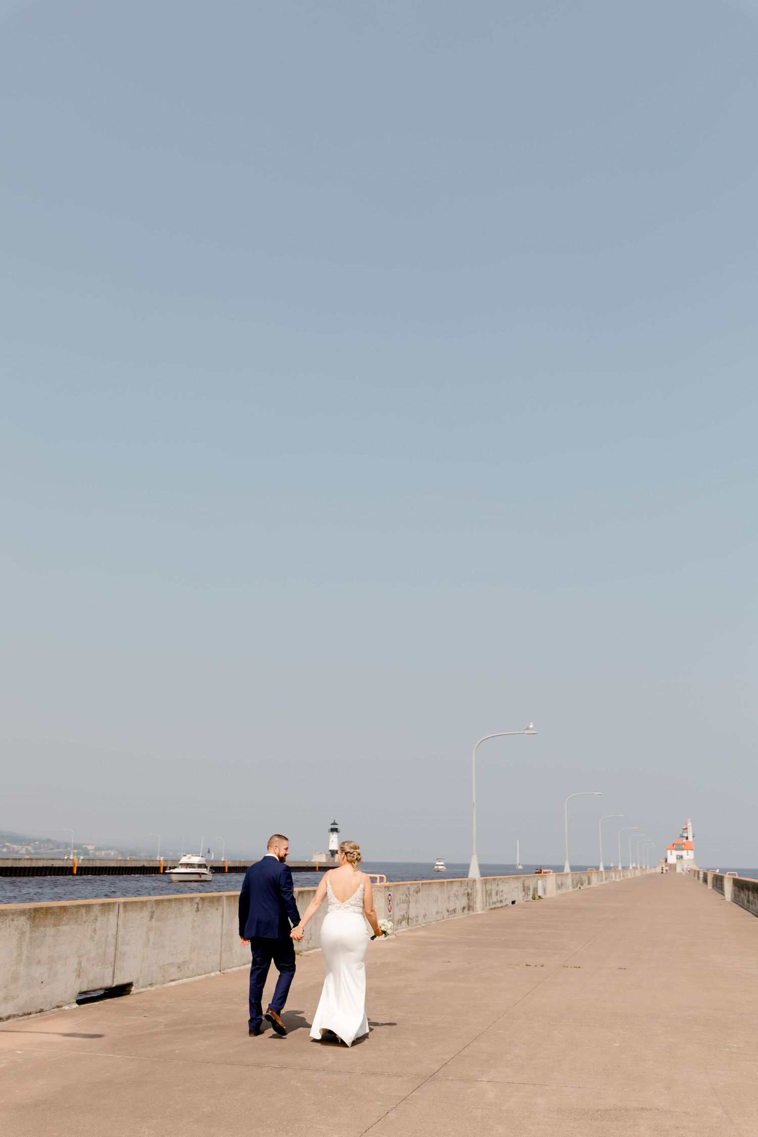 Bride and groom walking hand in hand on a waterfront pier with a lighthouse in the background on their wedding day.