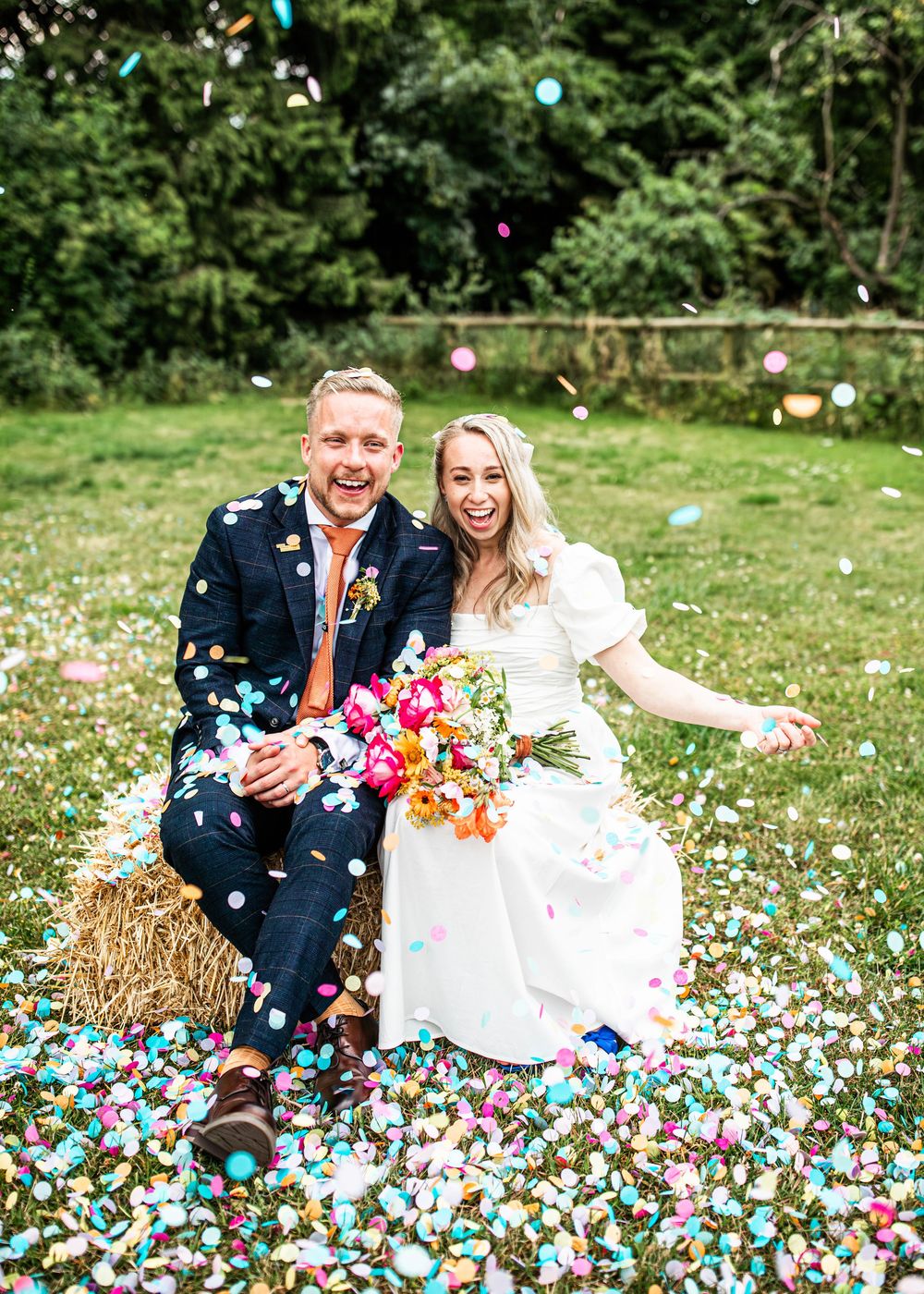 Joyful bride and groom sitting on hay bale surrounded by colorful confetti in a green meadow.