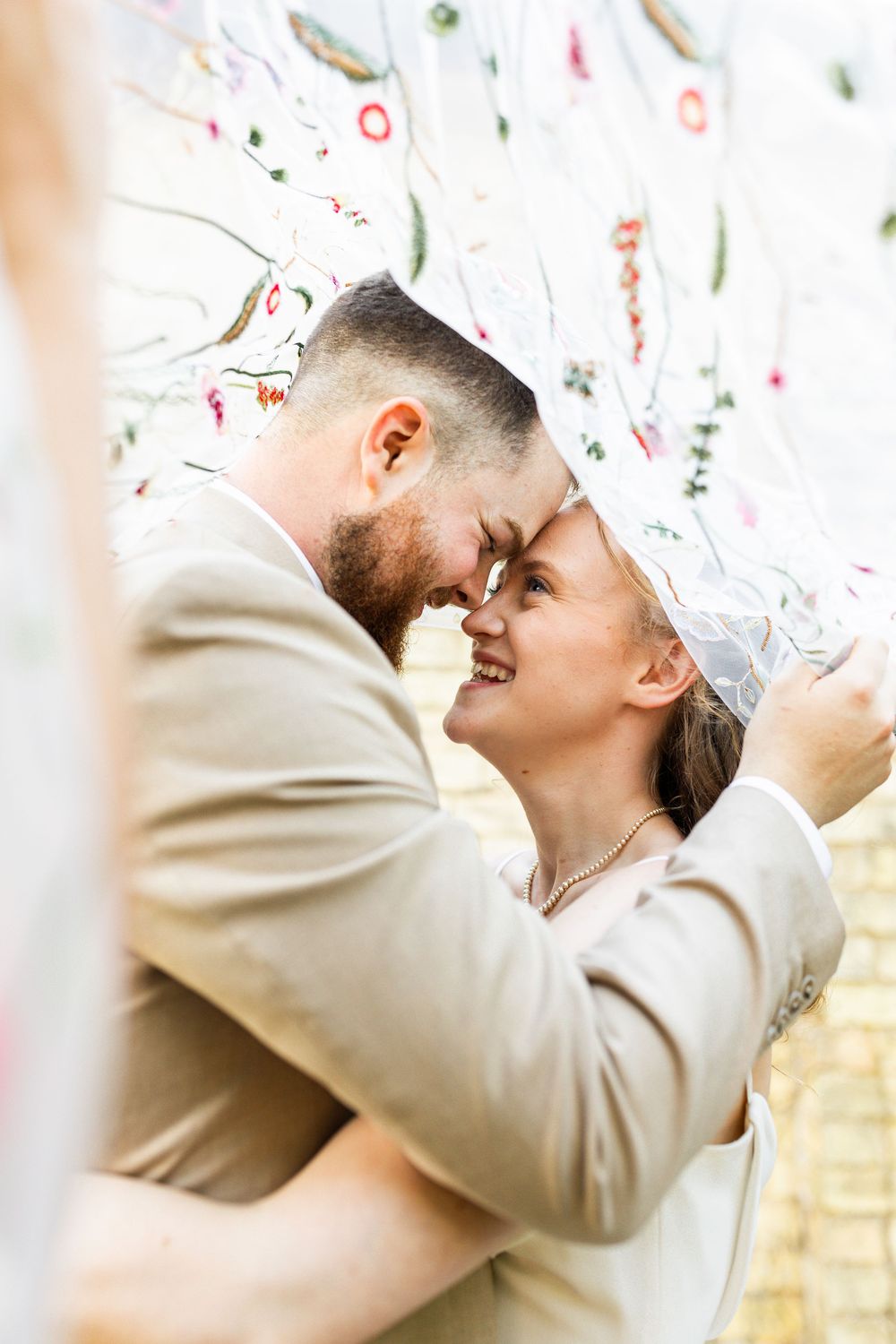 Romantic bride and groom sharing an intimate moment under a floral embroidered veil, foreheads touching.