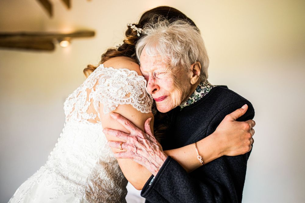 Bride in lace wedding dress sharing an emotional hug with elderly grandmother who is crying.