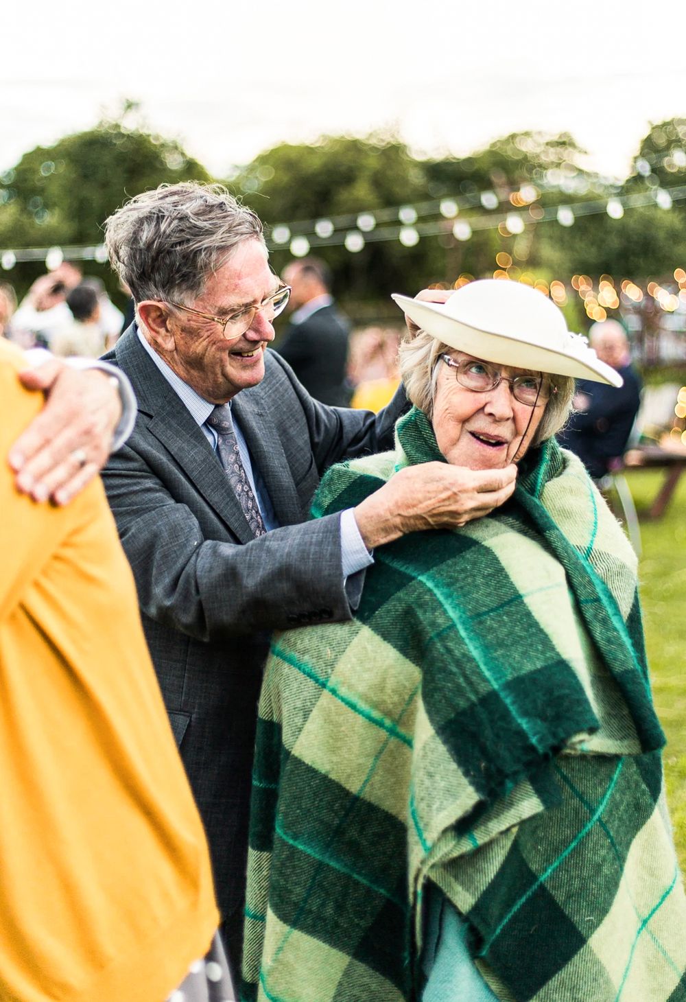 Elderly man wrapping a green plaid blanket around a woman at an outdoor wedding reception.