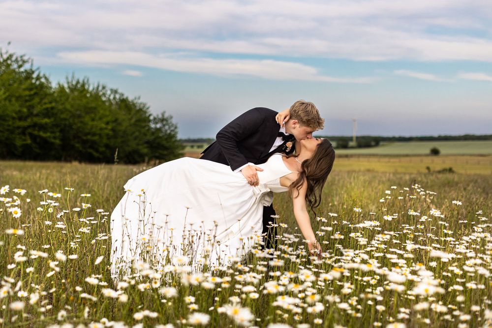 Bride and groom sharing romantic dip kiss in a field of white daisies on their wedding day.