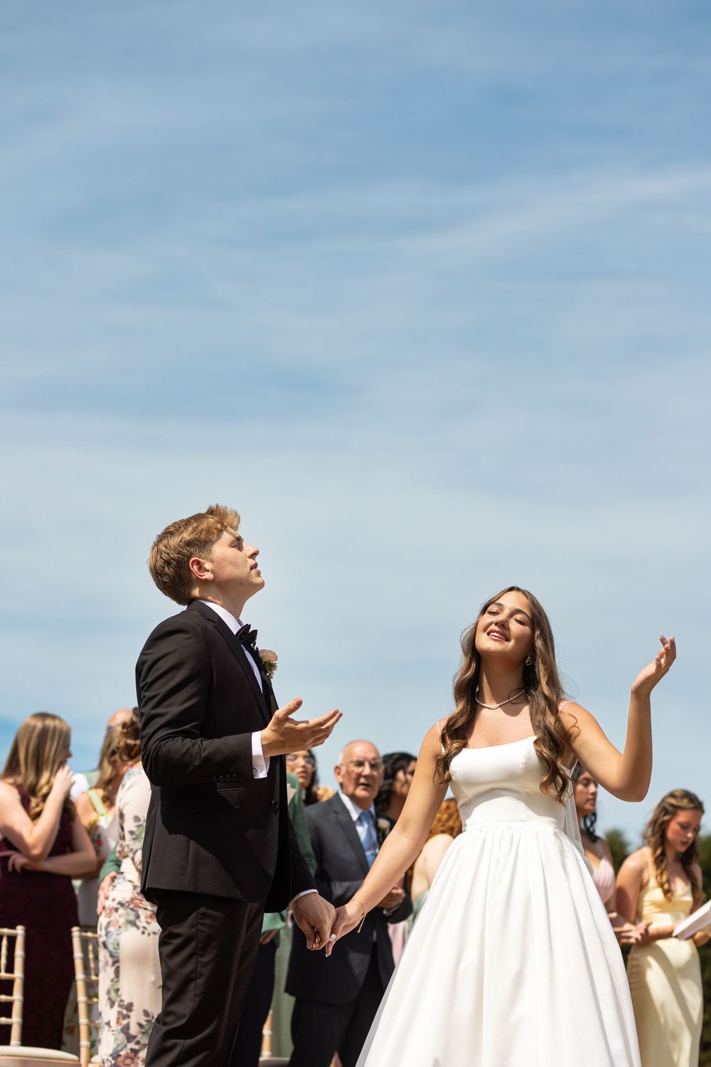 Bride and groom looking up joyfully at confetti during outdoor wedding ceremony under blue sky.