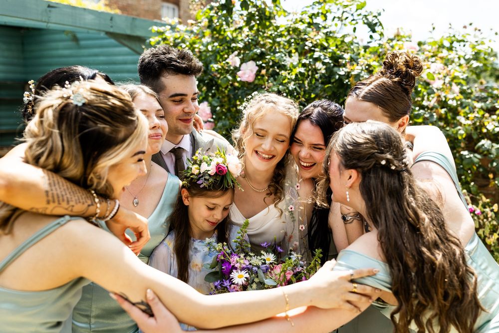 Bride and wedding party share a warm group hug outdoors in sunshine, bridesmaids in mint green dresses.