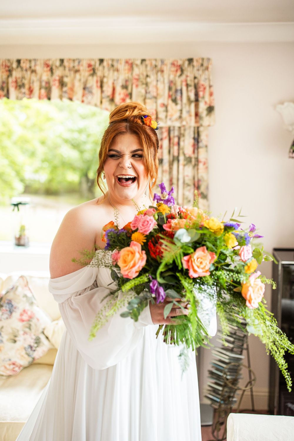 Joyful bride in white off-shoulder gown holding a vibrant colorful bouquet of roses and wildflowers indoors.