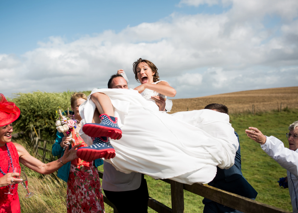 Groom lifts bride over a fence as laughing wedding guests help during outdoor celebration.