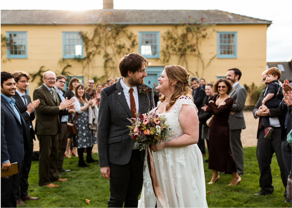 Newlyweds smile at each other as guests applaud during outdoor wedding reception ceremony.