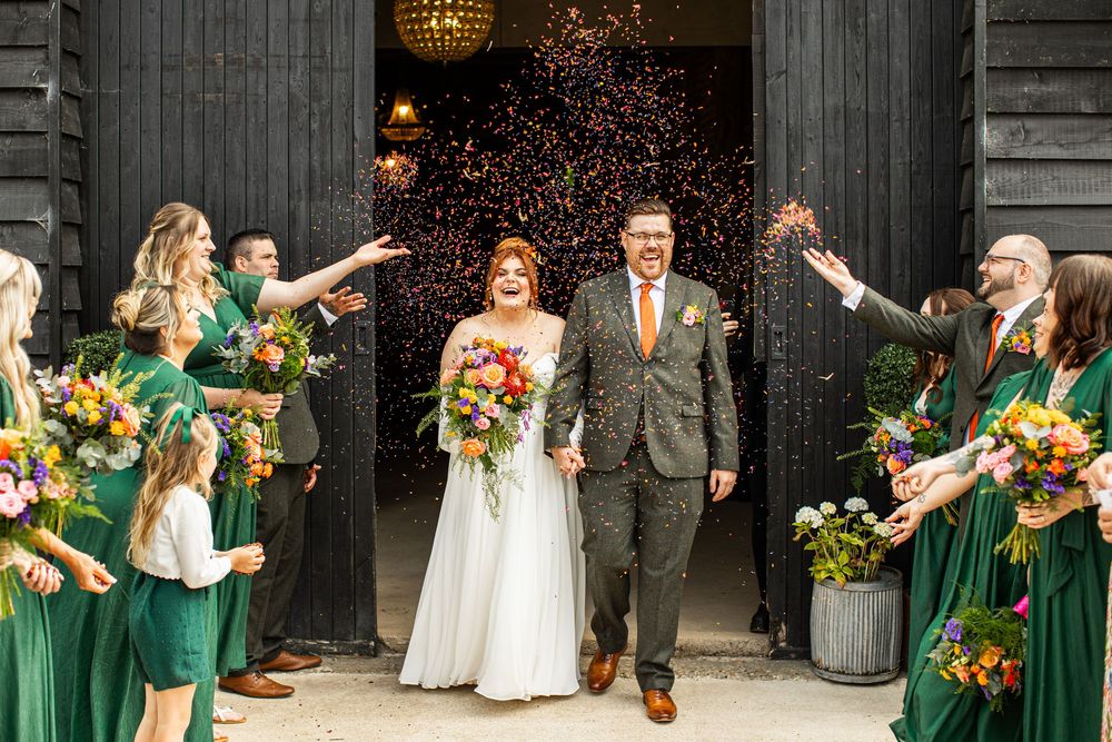 Newlyweds exit barn venue through confetti shower, bridesmaids in emerald green dresses celebrating around them.