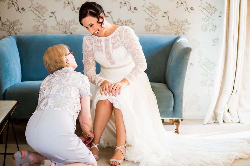 Mother kneeling to help bride put on wedding shoes beside a blue sofa in elegant room.