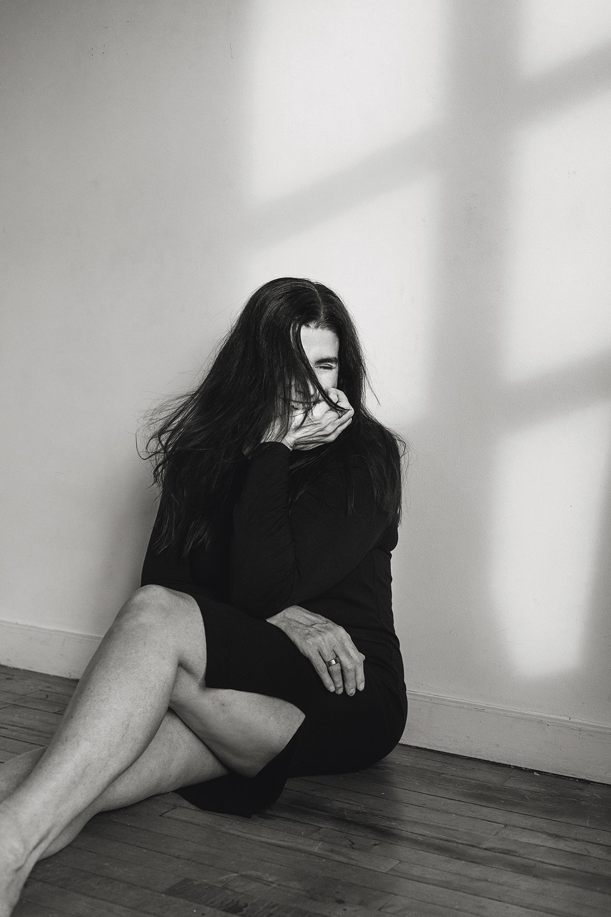 Black and white artistic photograph showing a person in dark clothing curled up against a wall with dramatic shadows.