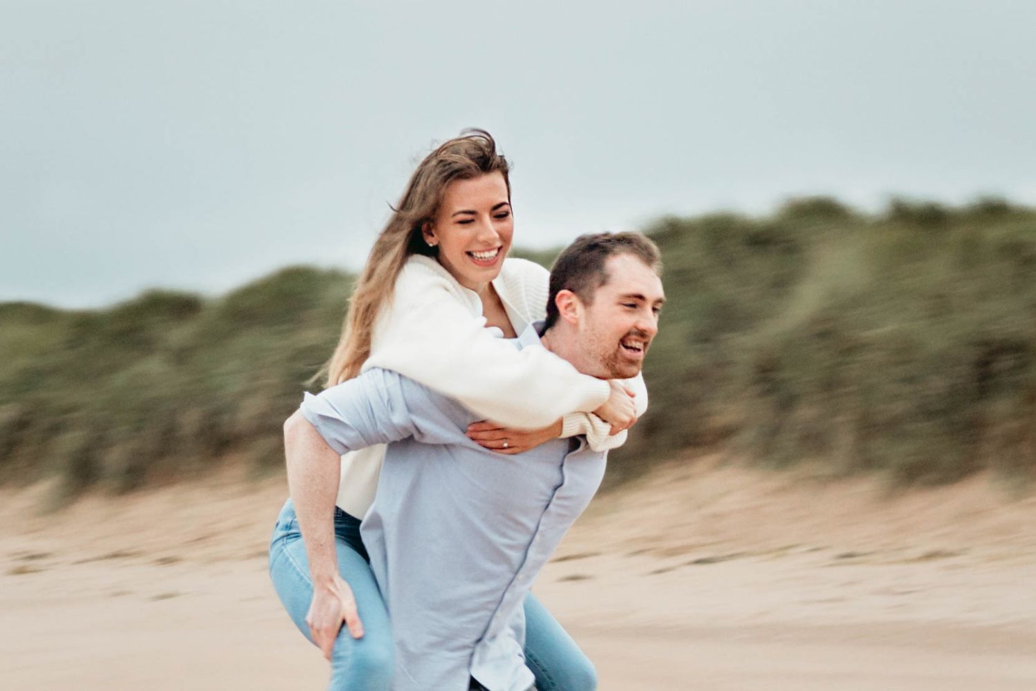 Man carries woman on his back as they run down a beach during a couple engagement photography session.