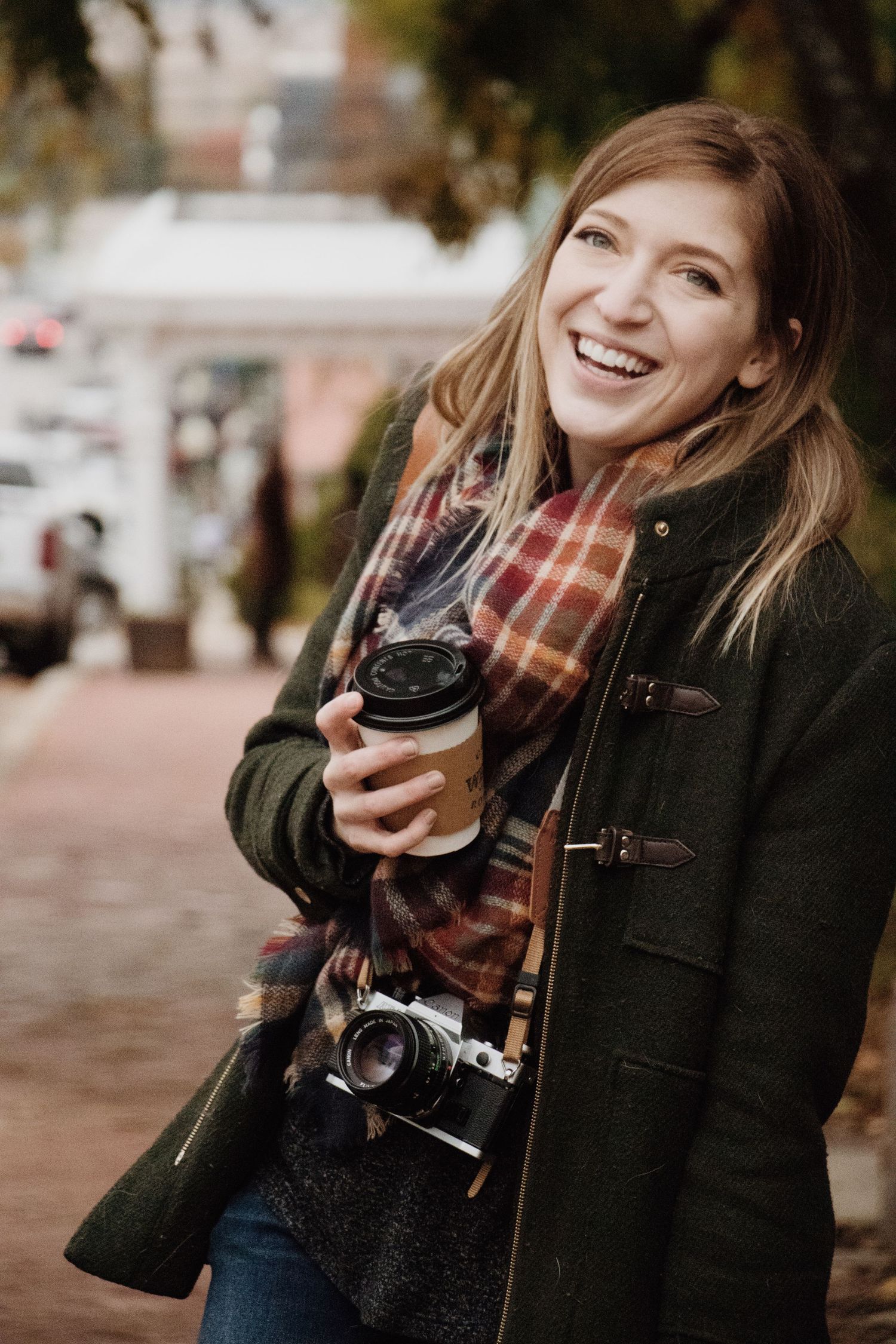 A Kansas City photographer wearing a coat and scarf holds a film camera and cup of coffee as she laughs and looks beyond the camera.