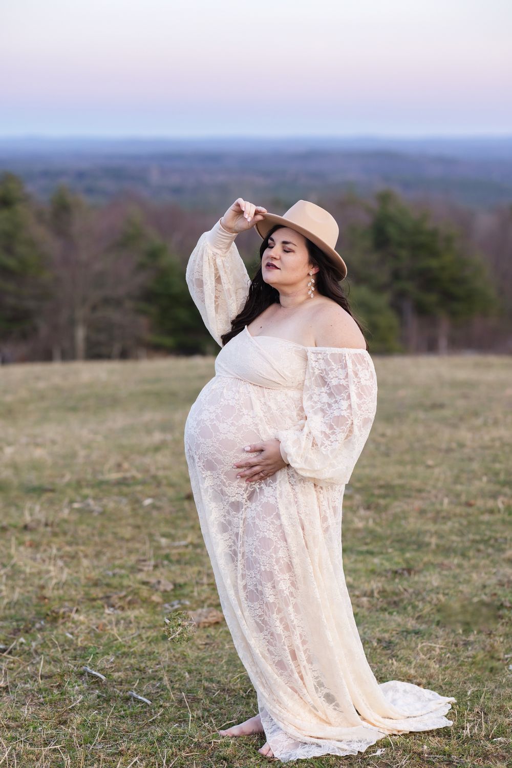 An expecting mother wears cream lace dress and beige velvet hat for maternity photos in Stoddard, NH at blue hour overlooking mountains.