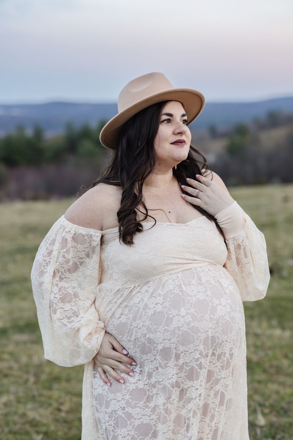 An expectant mother wears a cream lace dress and a beige velvet hat for maternity photos at a farm in the Lakes Region of New Hampshire.
