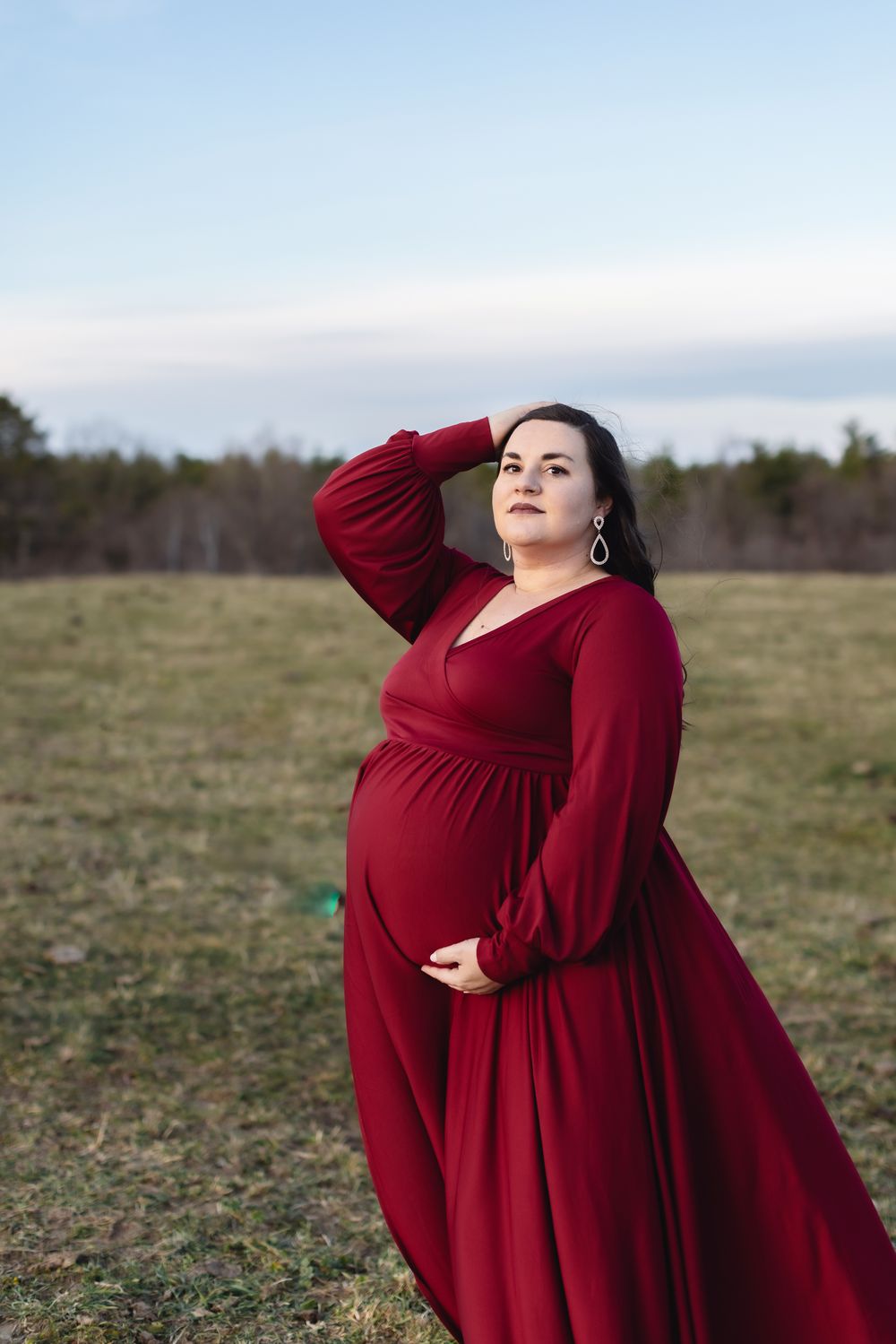 An expectant mother with dark brown hair wears a red gown for maternity photos at a farm near Concord, New Hampshire at sunset.
