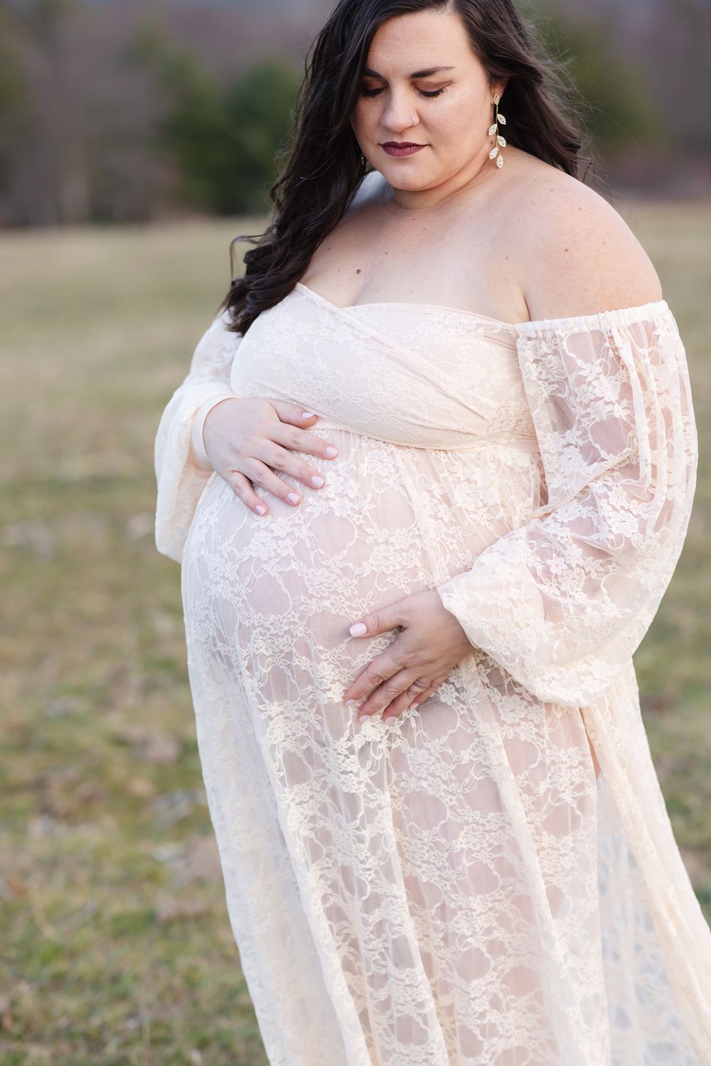 An expectant mother with brown hair wears a cream lace dress and looks down at her baby bump for maternity photos in a country field in NH.