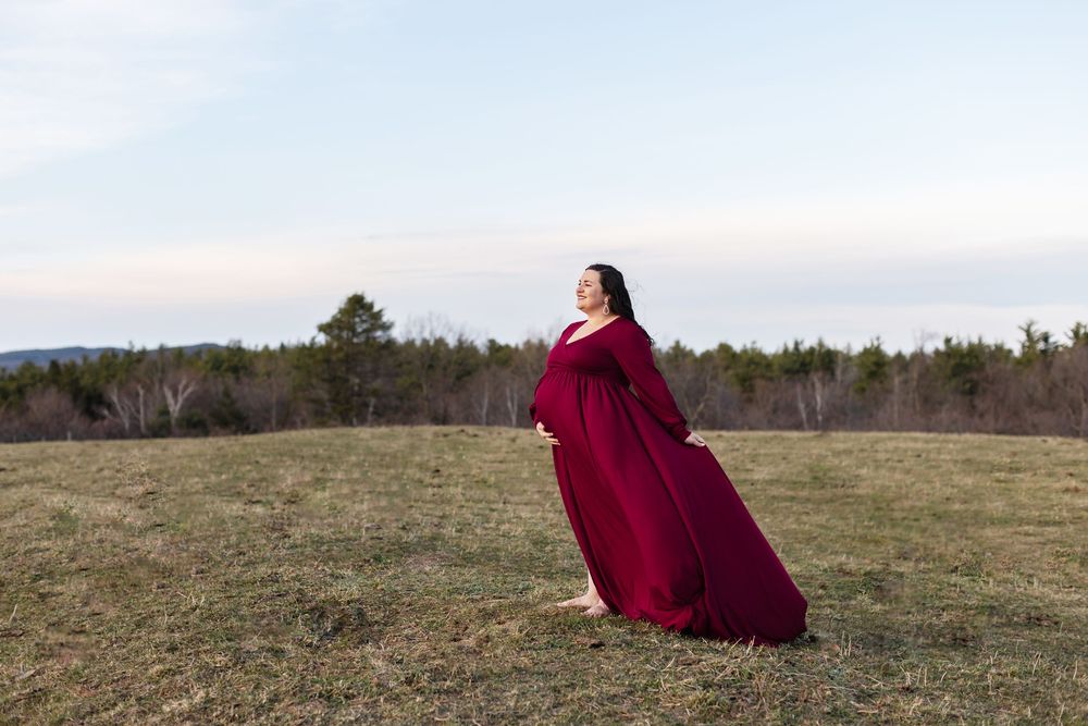 A happy expectant mother wears a burgundy gown for maternity photos in a country field at sunset at Pitcher Mountain Farm in Stoddard, NH.