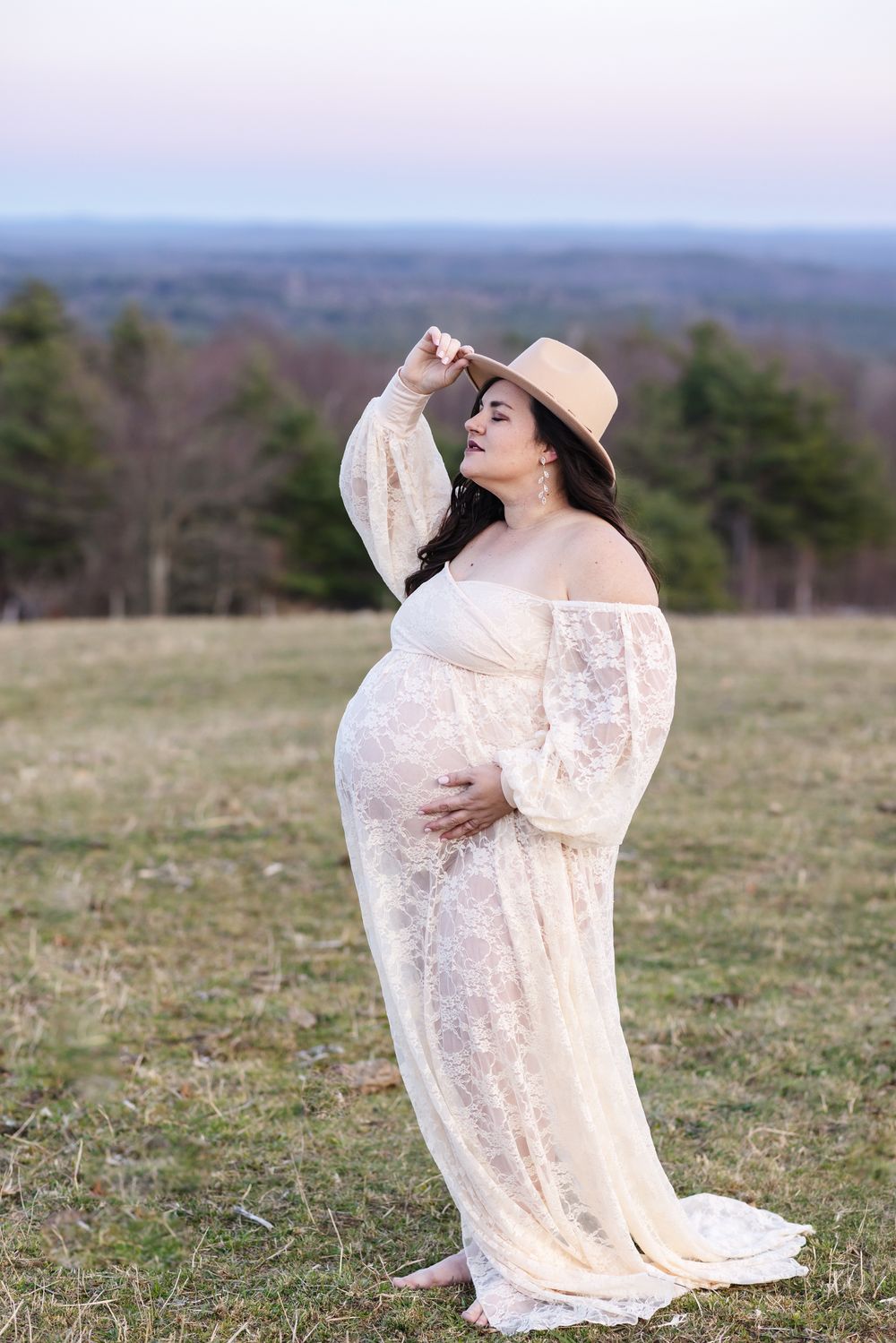 A pregnant mother tips her biege tan hat for maternity photos overlooking mountains at sunset at Pitcher Mountain Farm near Concord, NH.