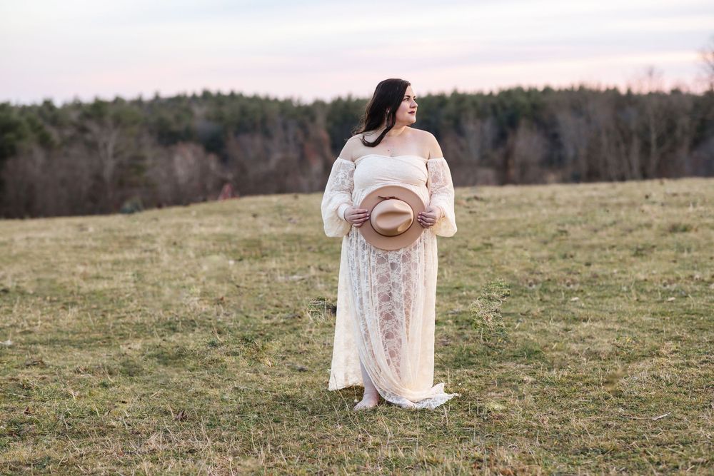 An expectant mother holds her beige hat over her baby bump for maternity photos at Pitcher Mountain Farm in Stoddard, New Hampshire.