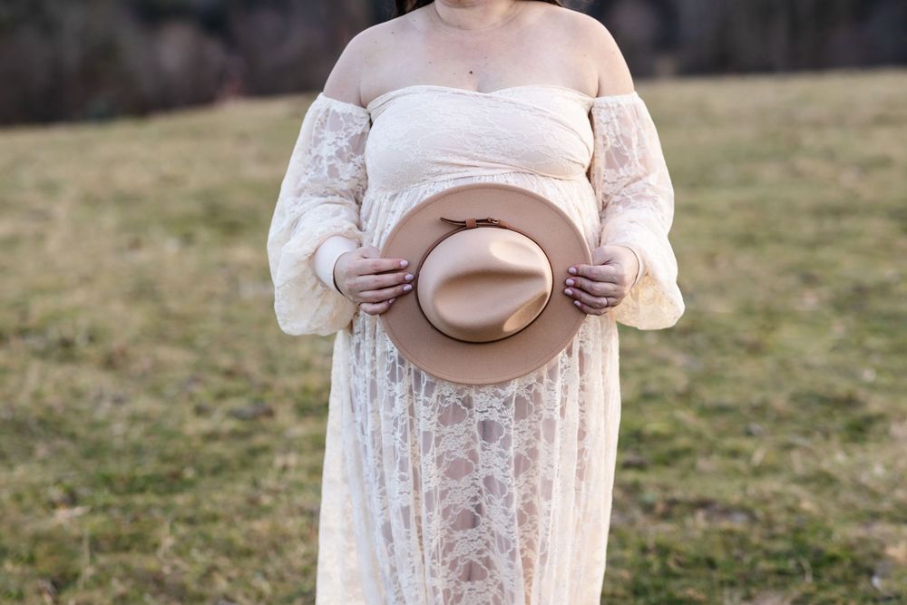 An expectant mother holds a beige hat over her baby bump for a close up photo at country photo shoot in New Hampshire.