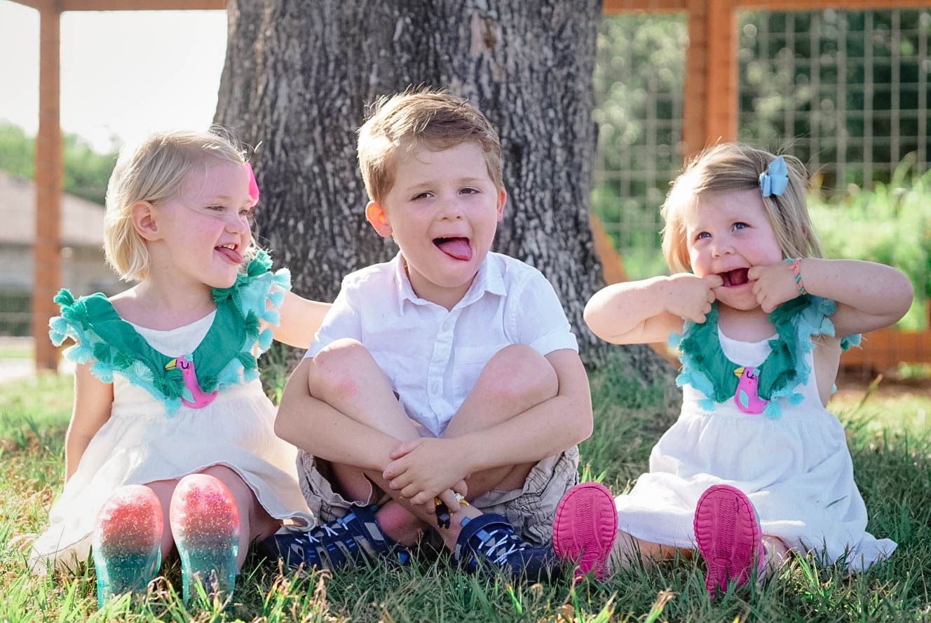 Young children sitting together on grass and laughing during an outdoor family photo session in Victoria, TX.