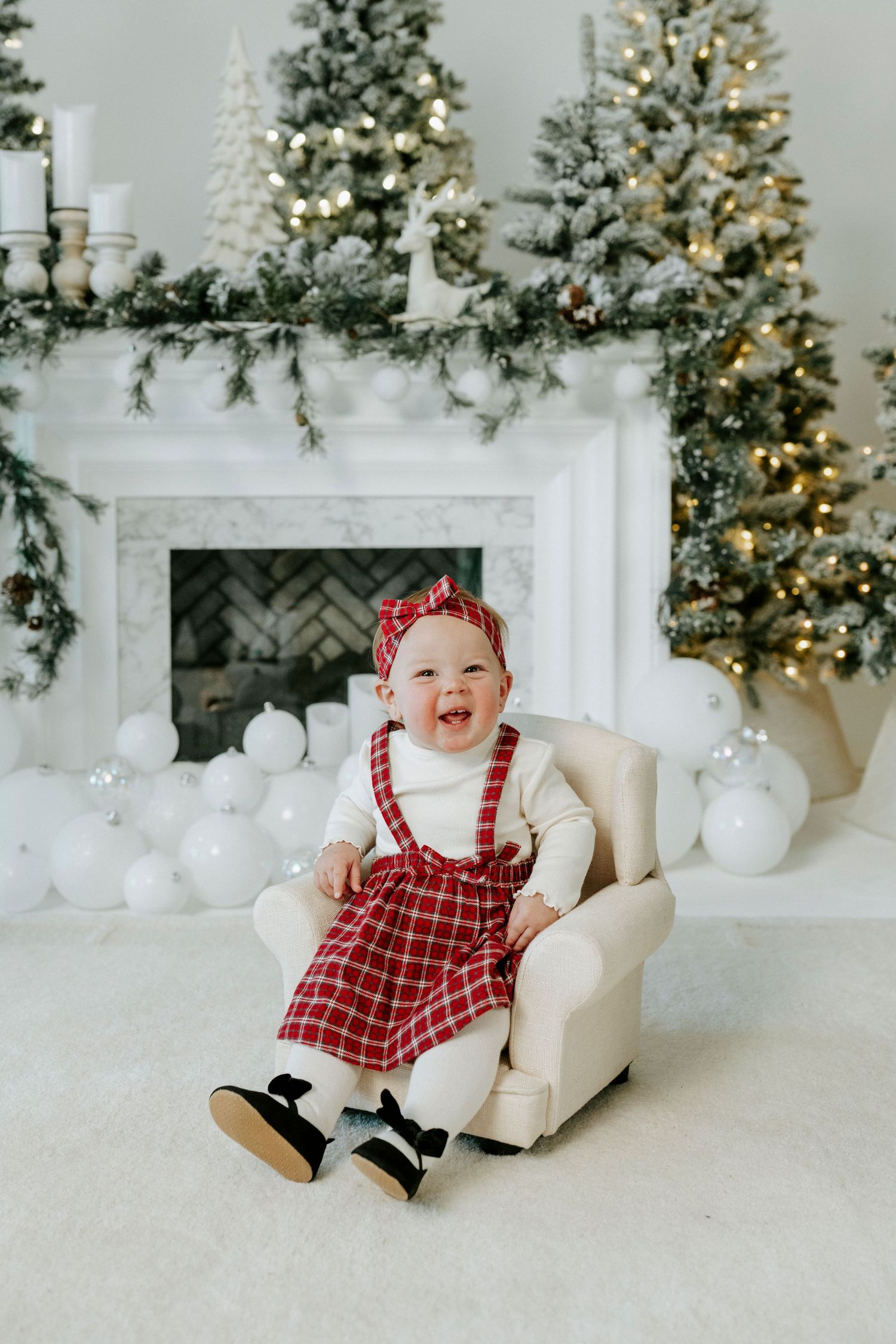 A baby in a red plaid holiday dress sits on a white chair with Christmas trees and decorations in the background.