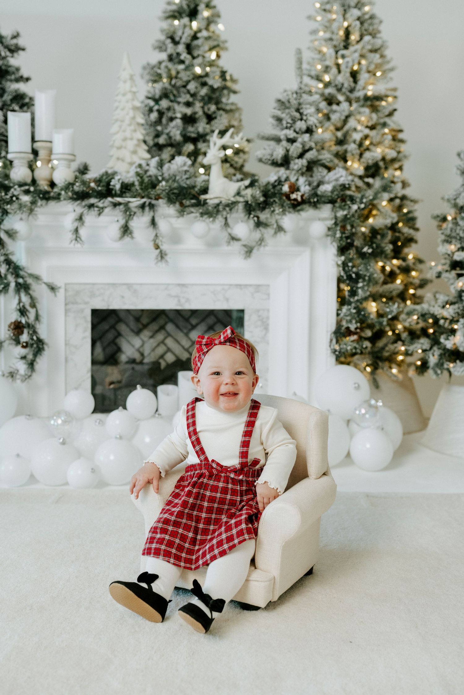 Baby in red plaid Christmas dress sits on white floor with decorated trees and fireplace in background.