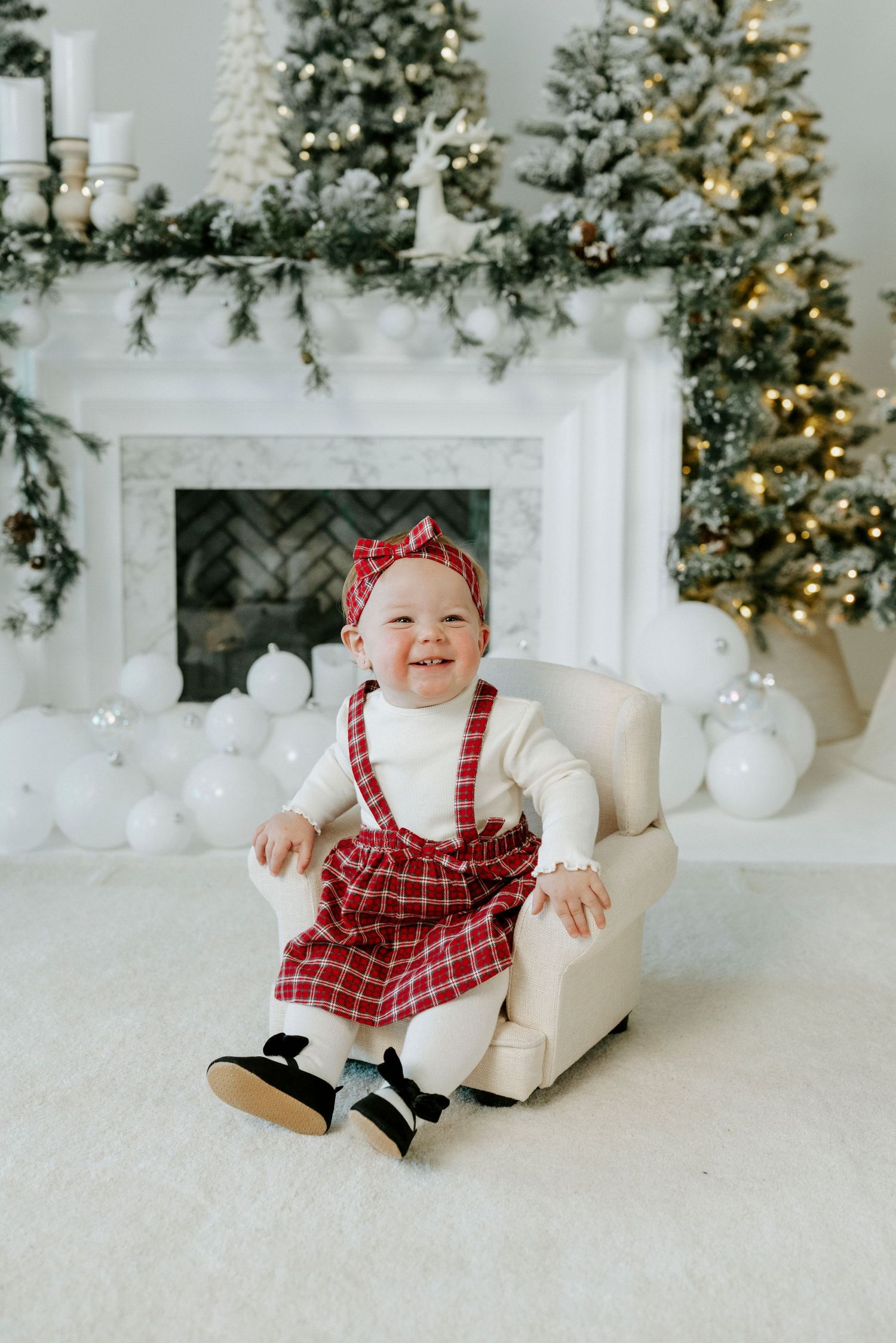 Festive holiday scene with decorated fireplace, Christmas trees, and small child in red plaid outfit sitting in white chair.